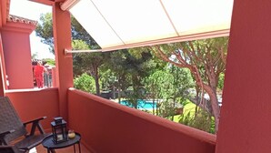 A balcony with a view of a swimming pool and trees. There are two chairs and a small table with a lamp.