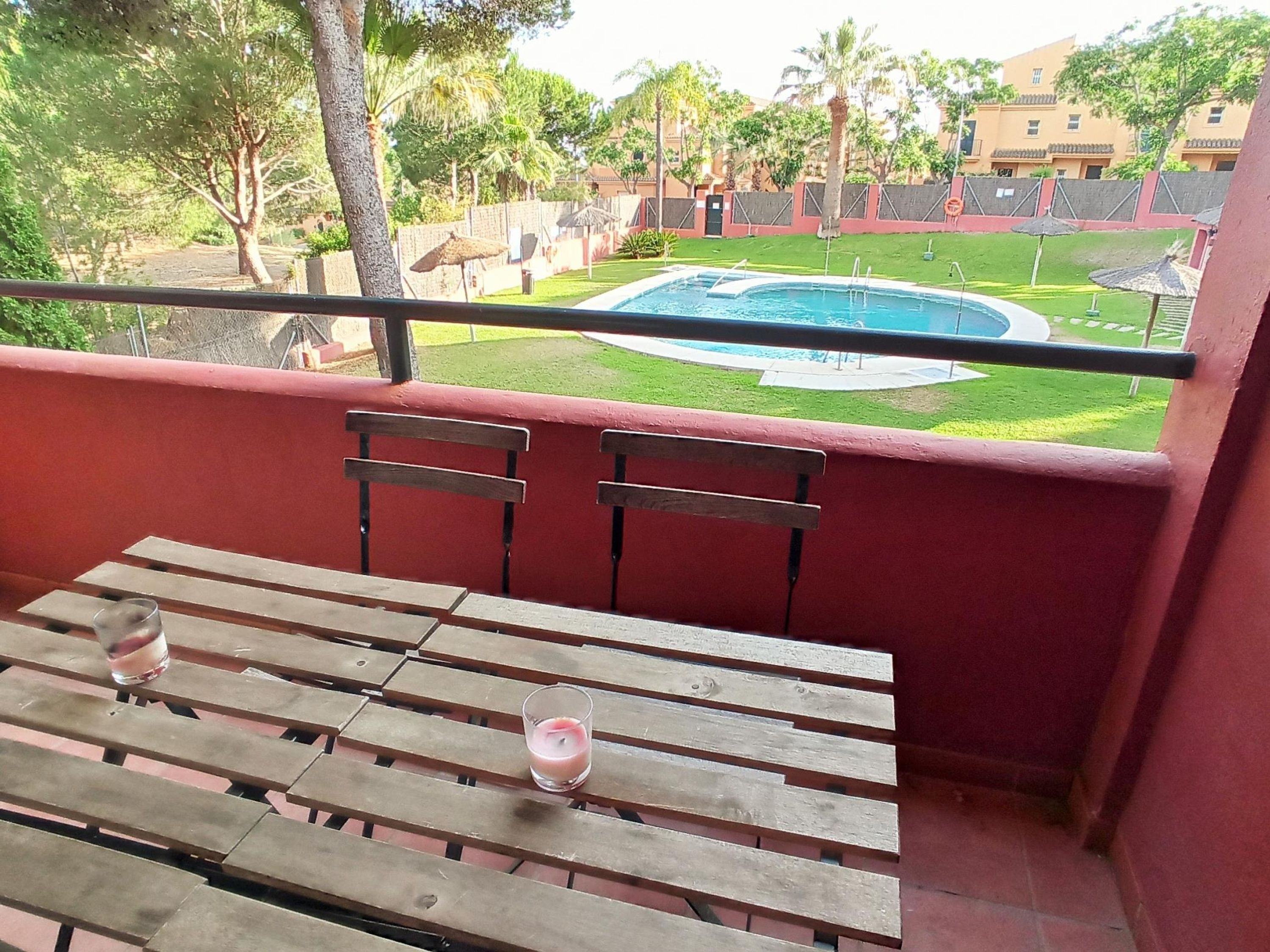 A balcony with a wooden table and chairs, overlooking a swimming pool and a well-maintained lawn.