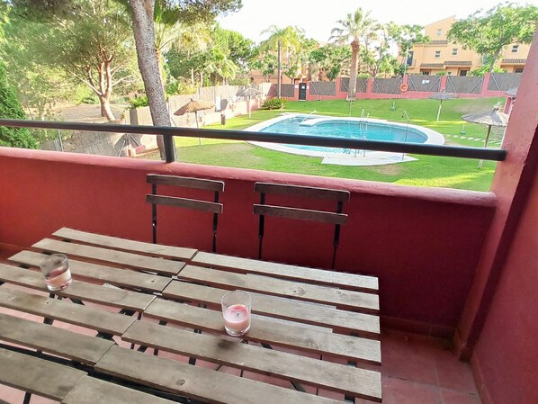 A balcony with a wooden table and chairs, overlooking a swimming pool and a well-maintained lawn.