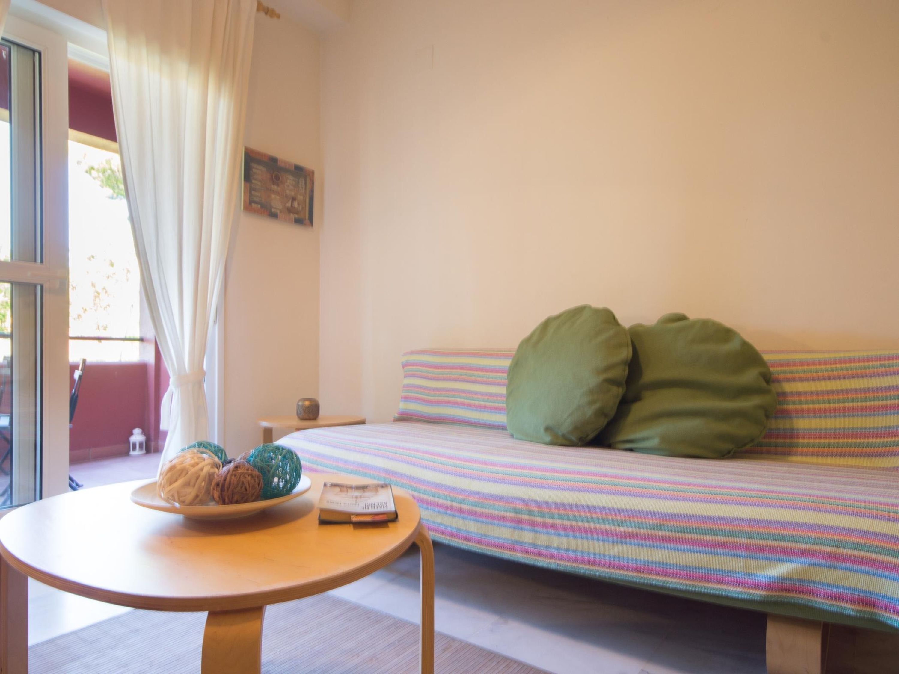 A room with a bed, a round table with a bowl of fruit and a book, and a window with curtains.
