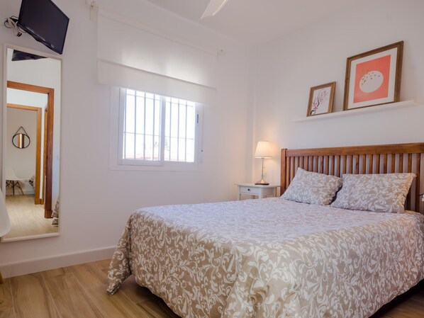Un dormitorio con una cama grande, una mesita de noche con lámpara, una ventana con rejas y un televisor colgado en la pared.