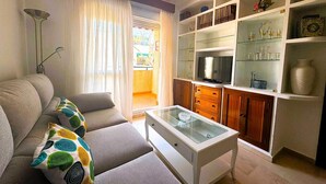 A living room with a sofa, a glass coffee table, a TV, and a bookshelf. - Apartamento Cabo Noval 15158 (Sanlucar de Barrameda)