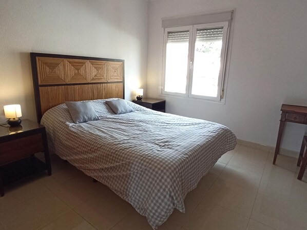 Una cama bien hecha con colcha a cuadros y almohadas, un cabecero de madera, dos mesitas de noche con lámparas y una ventana con persianas.