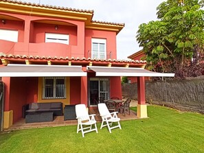 Una casa de dos pisos con fachada roja, un porche cubierto con mesa y sillas, y un césped bien cuidado.