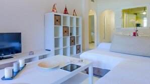 A modern hotel room with a flat-screen TV, a white console table with a tablet, a white sofa, a white cabinet with decorative items, and a large mirror.
