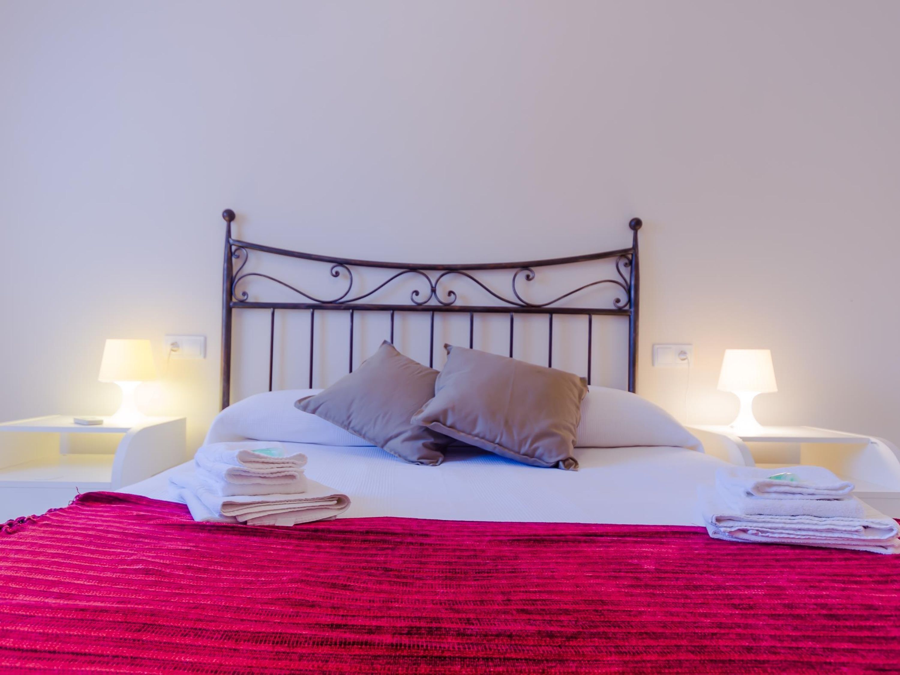 A neatly made bed with a red blanket, two pillows, and a metal headboard. There are two bedside tables with lamps on either side of the bed.