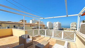 Una terraza en la azotea con mobiliario de mimbre, vista al mar y edificios circundantes.