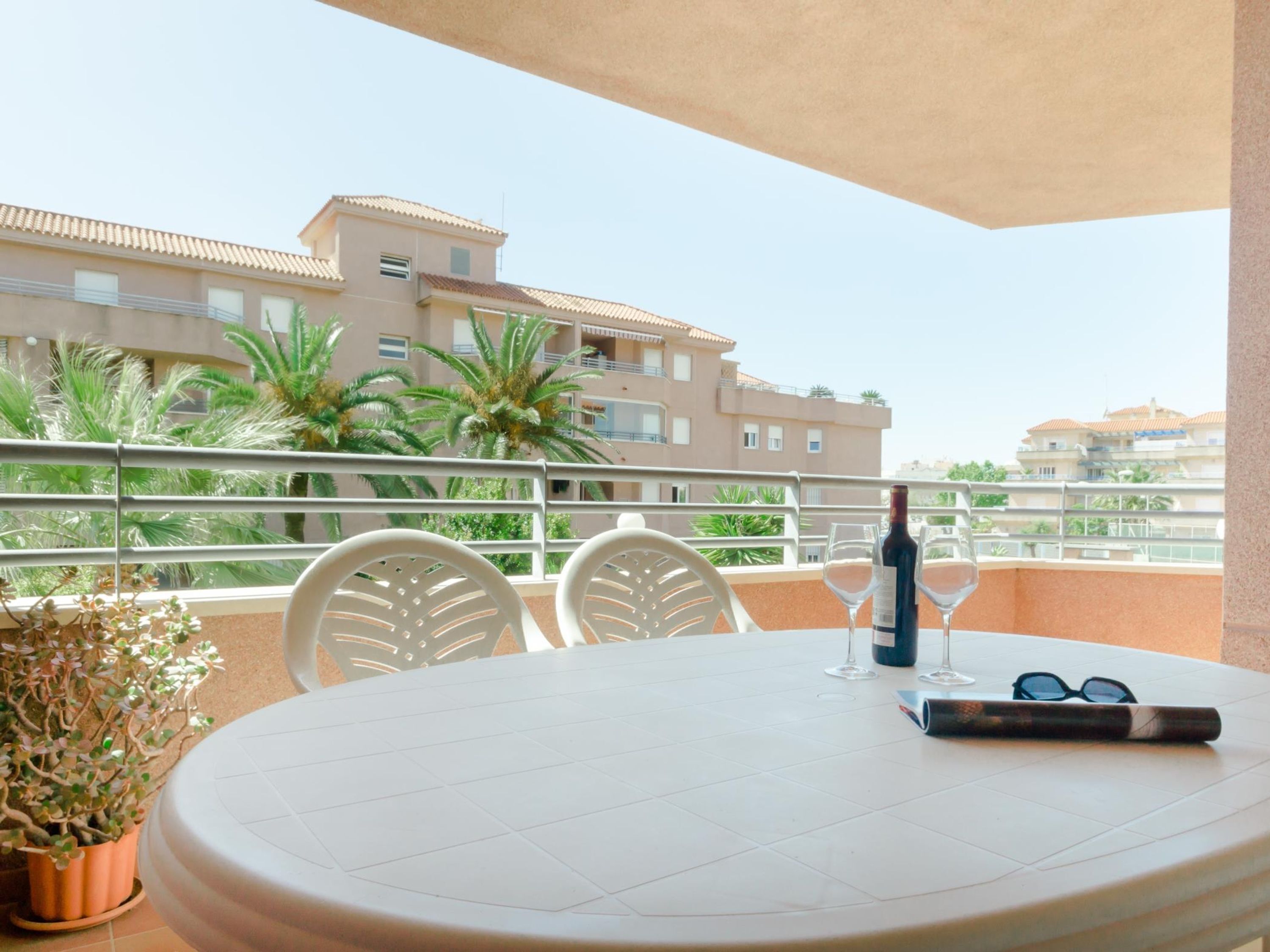 A round table with two chairs, a bottle of wine, and a pair of glasses on a balcony overlooking a residential area with palm trees and buildings.
