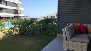 A balcony with a wicker sofa, a pool, and a view of modern buildings. - Bah a Blanca II 18923 (El Puerto de Santa Maria)