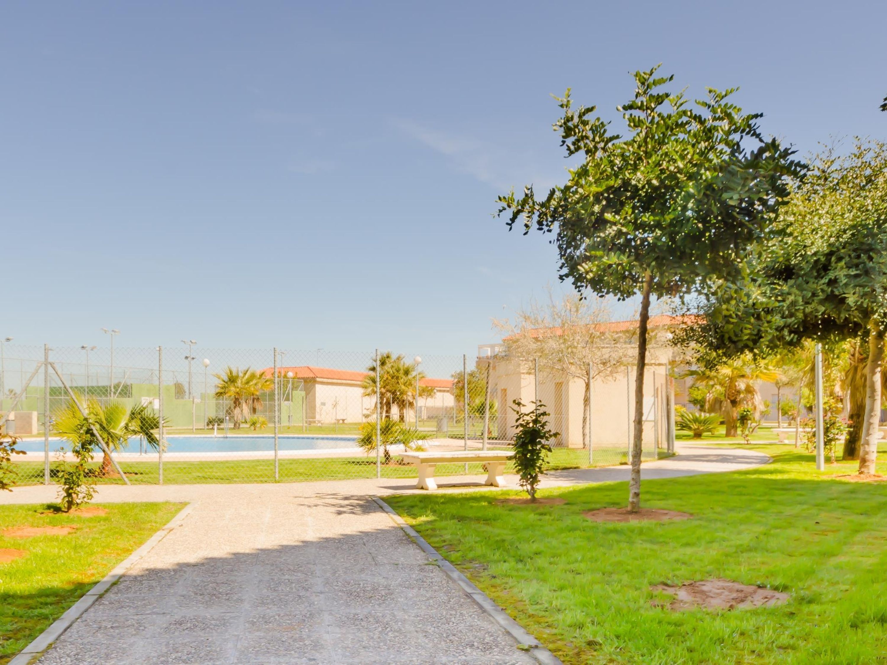 A pathway leading to a pool area with a fence, palm trees, and a building with a red roof.