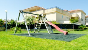 Un terrain de jeux avec des balançoires et un toboggan, situé devant un immeuble doté de balcons.