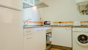 Een kleine keuken met witte kastjes, een inbouwmagnetron, een wasmachine en een betegelde achterwand.