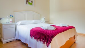 Een net opgemaakt bed met witte lakens en een roze deken, een nachtkastje met een lamp, en een aan de muur hangend schilderij.