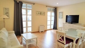 A living room with a white sofa, a small white table, a glass-top dining table with white chairs, a flat-screen TV on a stand, and large windows with curtains. - Ribera E 13489 in El Puerto de Santa Mar a (El Puerto de Santa Maria)