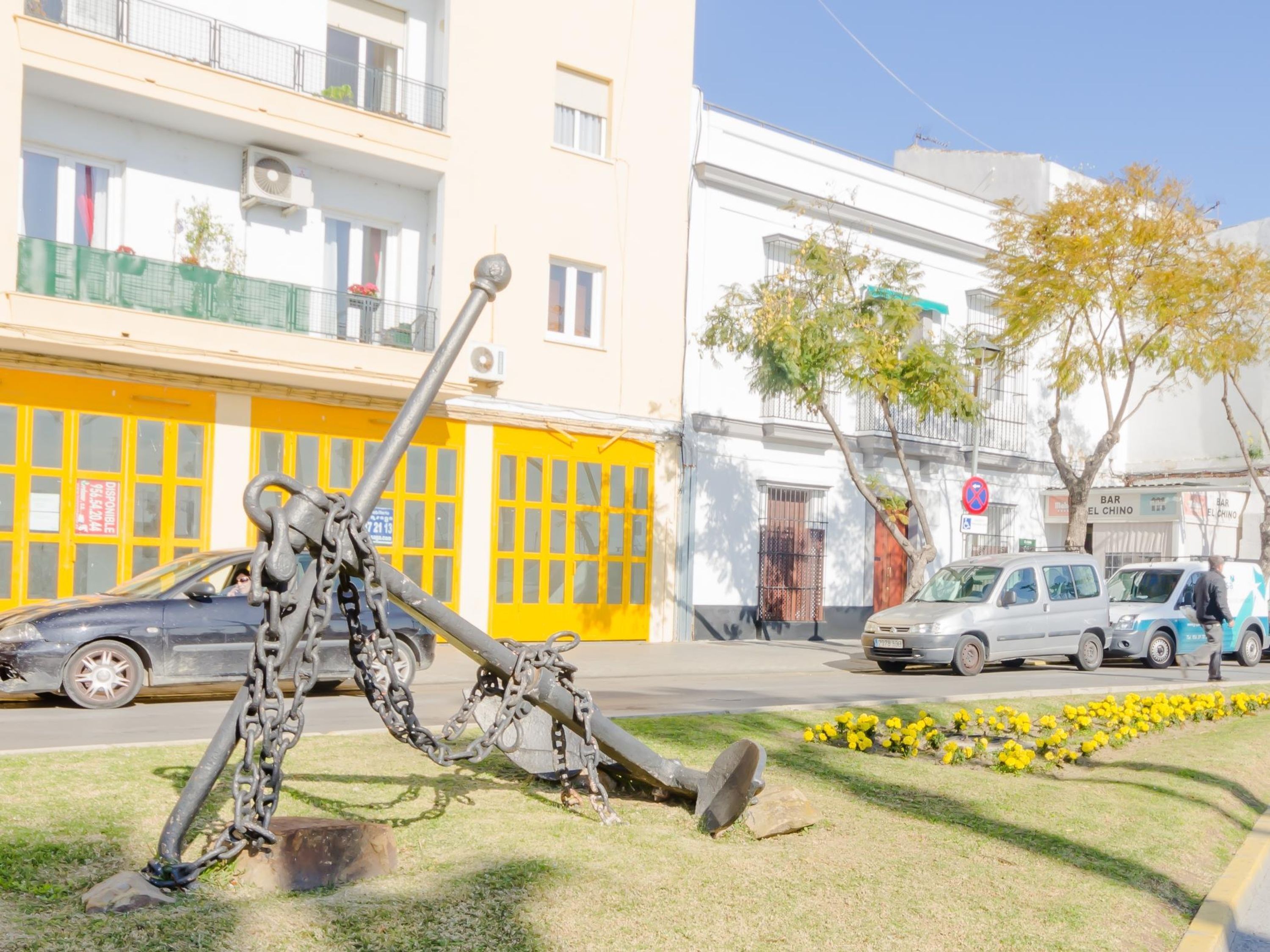 A street scene with a large metal sculpture in the foreground, parked cars, and buildings with balconies and windows.