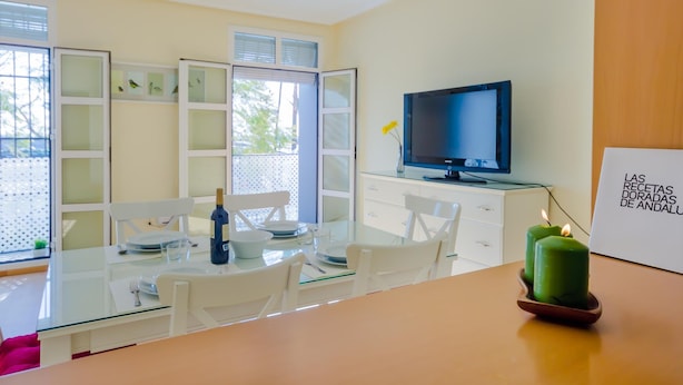 A modern dining area with a glass table, white chairs, a television on a stand, and a candle on the table. - Ribera E 13489 in El Puerto de Santa Mar a (El Puerto de Santa Maria)