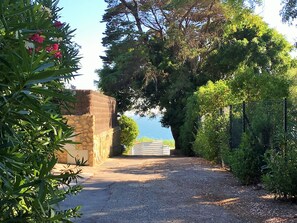 Un chemin de gravier mène à un mur de pierre surmonté d’une porte, le tout étant entouré d’une végétation luxuriante et d’arbres.