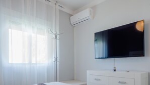 Un dormitorio moderno con cama, televisor montado en la pared, cómoda blanca y ventana con cortinas translúcidas.