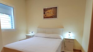 Ein ordentlich bezogenes Bett mit weißer Bettwäsche, zwei Nachttische mit Lampen, ein Fenster mit Jalousien und ein Gemälde an der Wand.