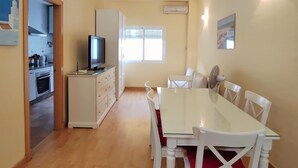 Ein Essbereich mit weißem Tisch und Stühlen, einem Fernseher auf einem Schrank und einer durchgehenden Tür erkennbaren Küche.