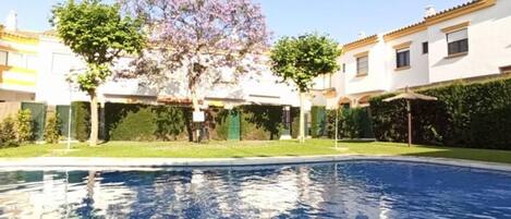 Una piscina con superficie de agua azul cristalina, rodeada por un césped cuidado y un edificio con tejado de baldosas.