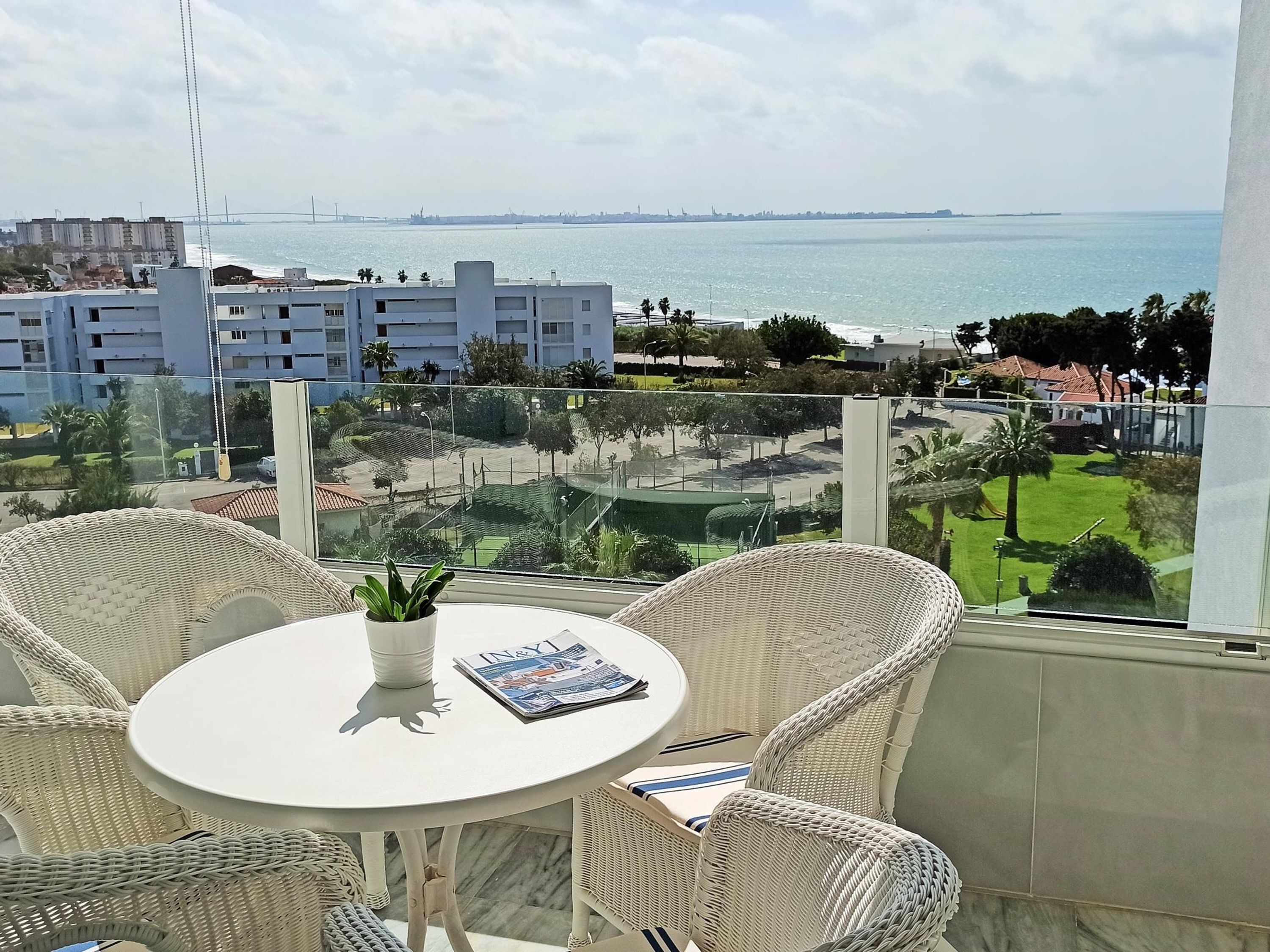 A balcony with wicker chairs and a table, overlooking a coastal view with buildings and a bridge in the distance.