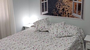 A bedroom with a bed, a nightstand, a window with a beach view, and a curtain. - Apartaclub 14116 in Chiclana (Chiclana de la Frontera)