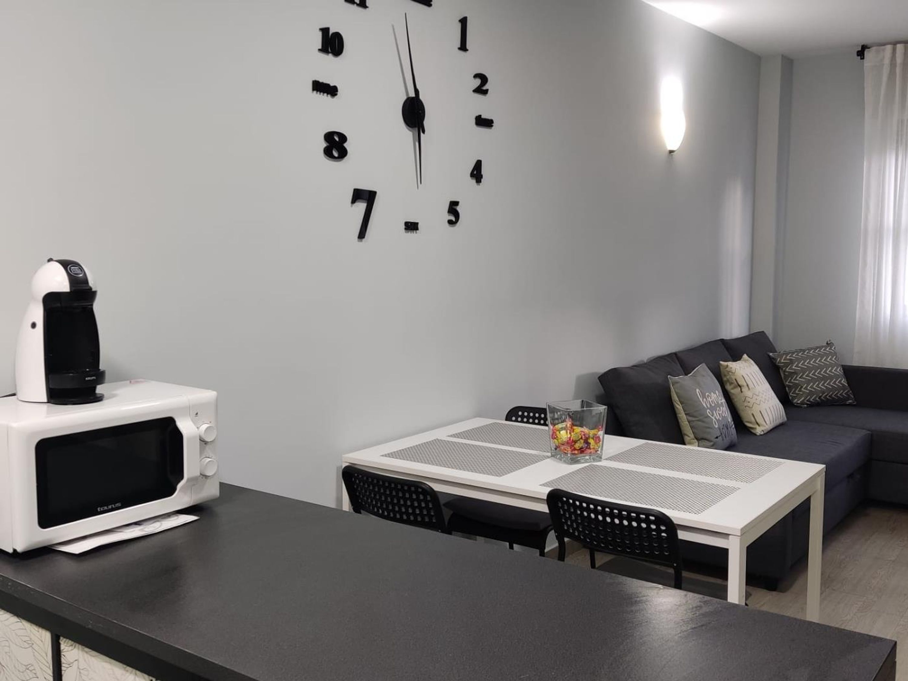 Una sala moderna con un reloj de pared blanco, una cafetera blanca, un sofá gris y una pequeña mesa con sillas.