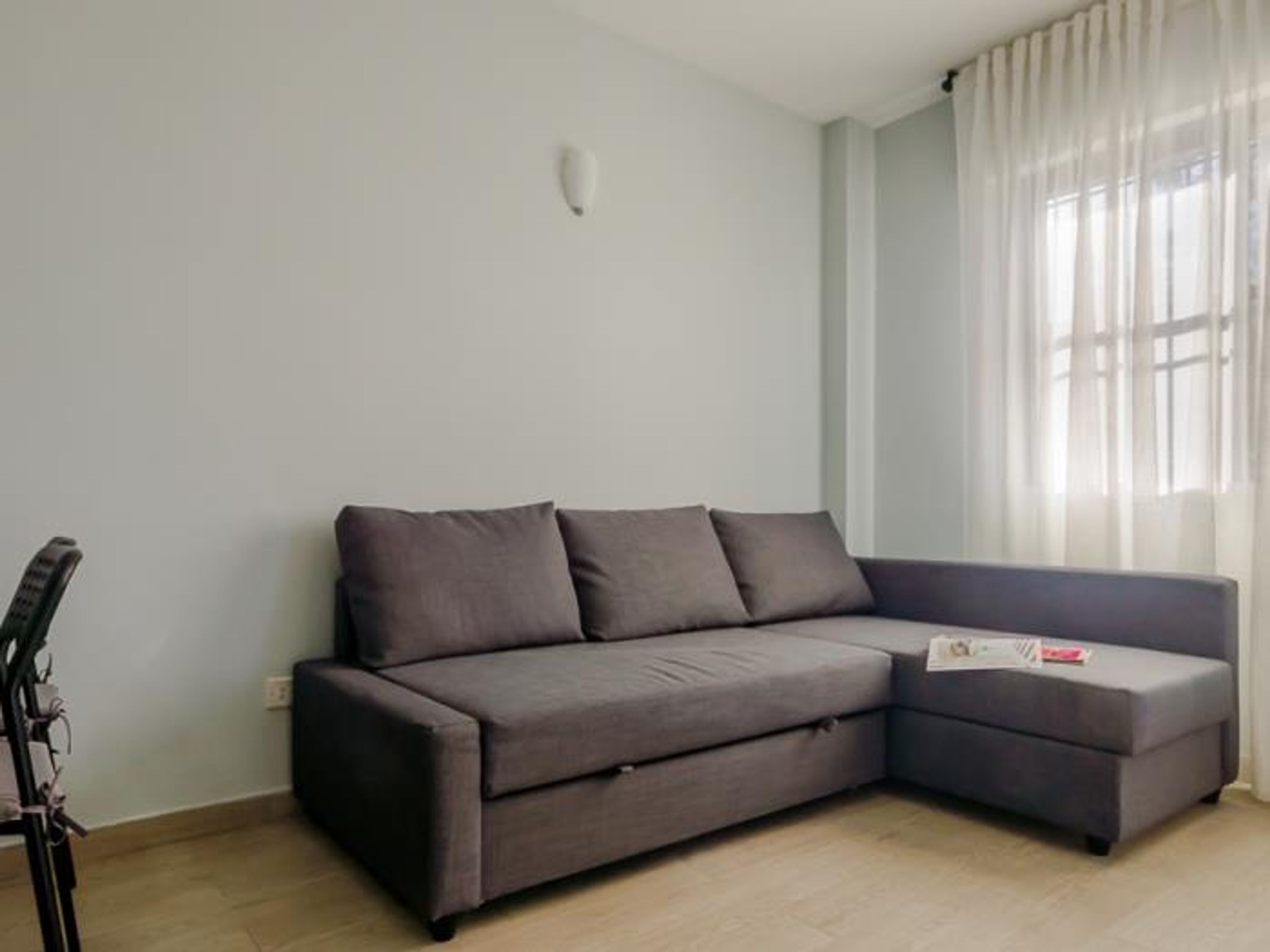 Un sofá seccional gris en una habitación con paredes de colores claros y una ventana con cortinas traslúcidas.