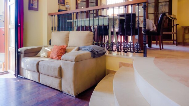 A living room with a beige sofa, a staircase with a glass railing, and a chandelier. - Villa Las Palomas 14988 (Chiclana de la Frontera)