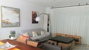 A modern living room with a dining area, grey sofa, wicker chairs, a wooden dining table, and a large painting on the wall. - Villa Laura 24952 in Conil de la Frontera (Conil de la Frontera)