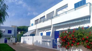 A white building with blue shutters, a small garden with red flowers, and a clear sky. - La Almadraba A7 147 in Chiclana (Chiclana de la Frontera)