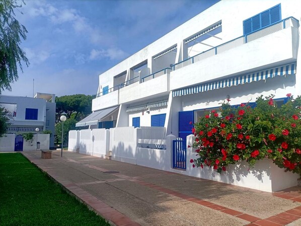 A white building with blue shutters, a small garden with red flowers, and a clear sky. - La Almadraba A7 147 in Chiclana (Chiclana de la Frontera)