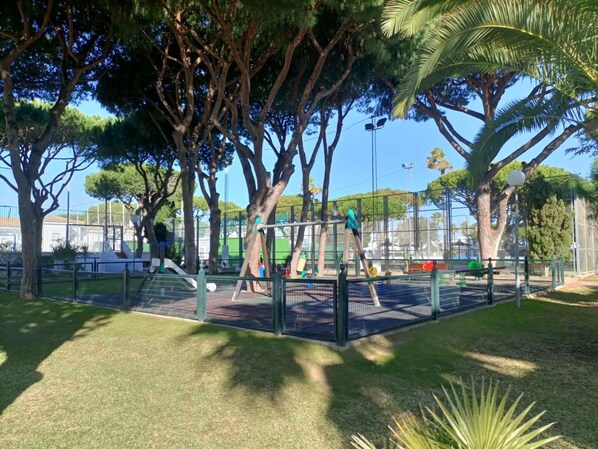 A playground with swings and climbing structures, surrounded by trees and a fence. - La Almadraba A7 147 in Chiclana (Chiclana de la Frontera)
