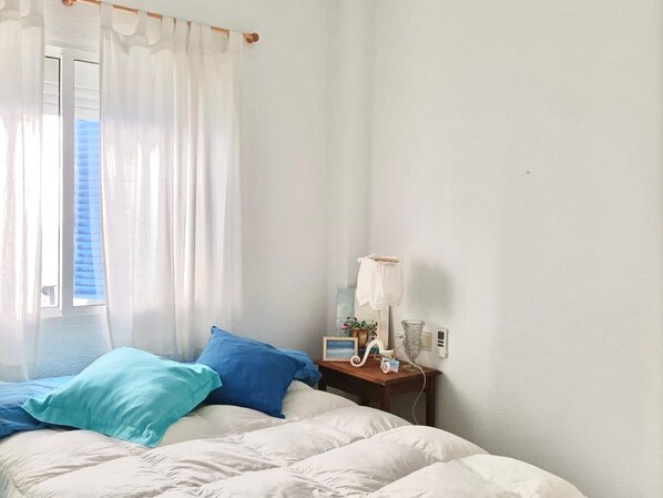 Un dormitorio con cama, mesita de noche con lámpara y ventana con cortinas.