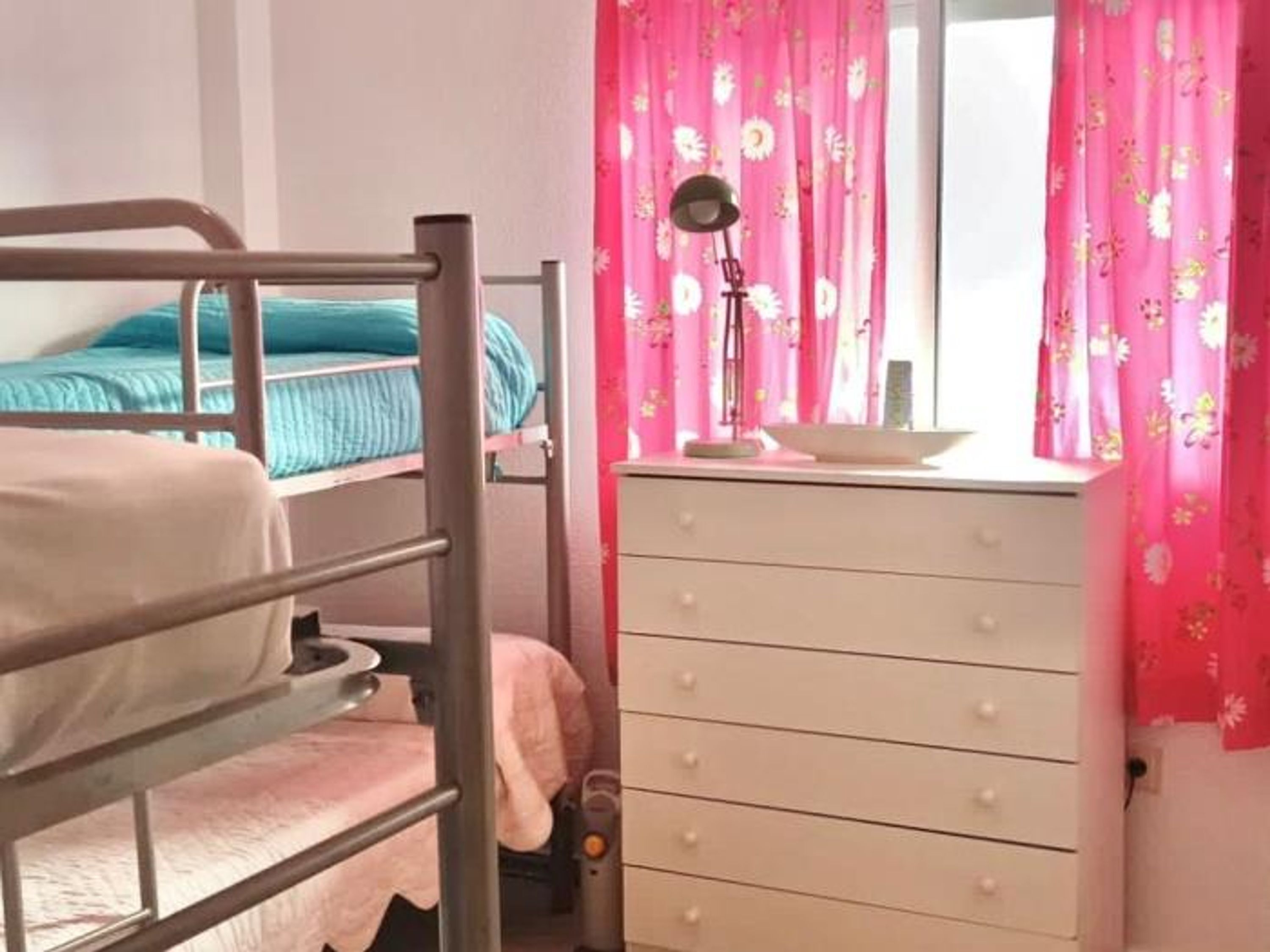 A bunk bed with a blue mattress, a white dresser with drawers, and pink floral curtains.