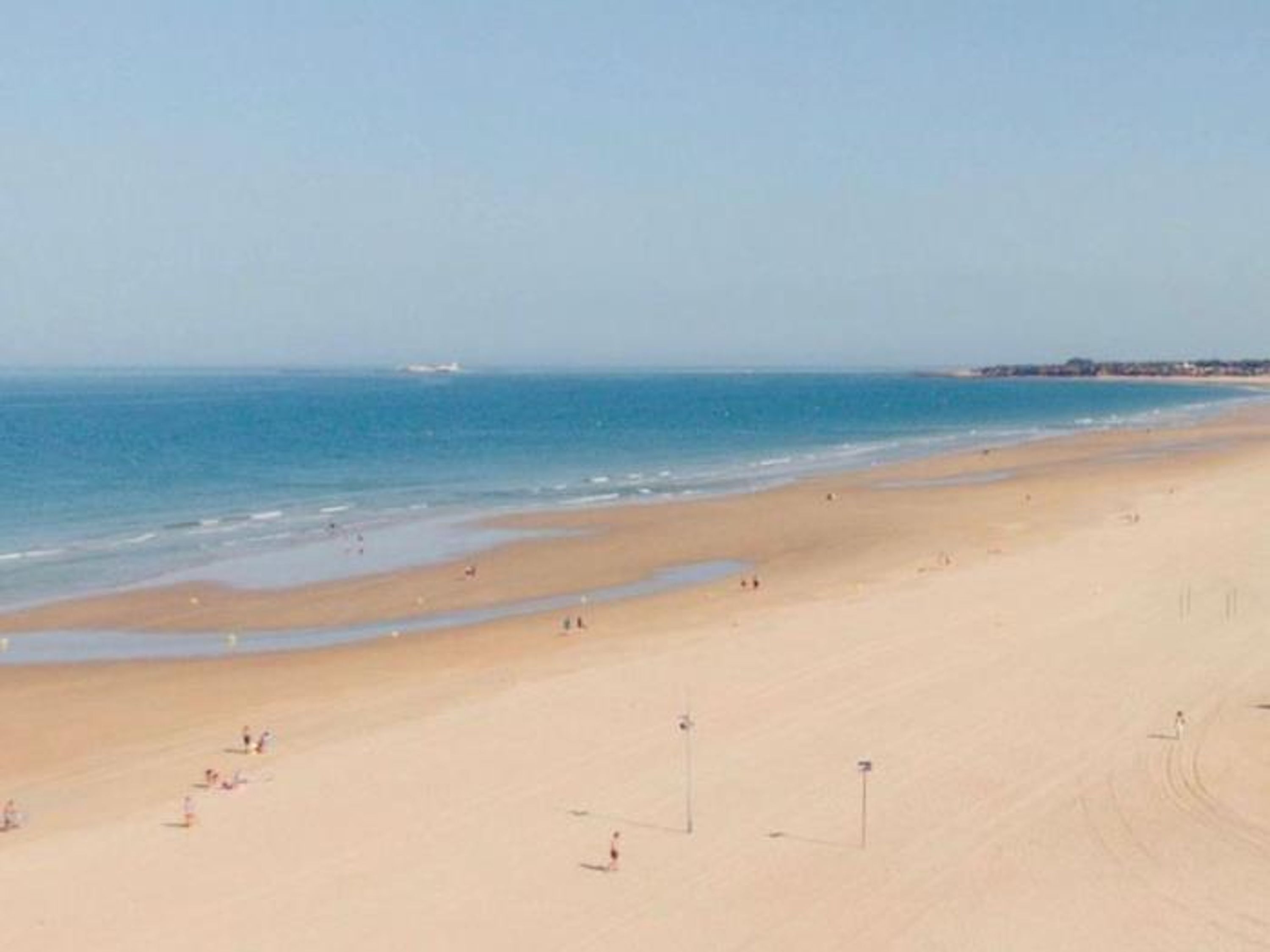 Una amplia playa de arena con un mar azul y transparente, algunas personas y edificios a lo lejos.