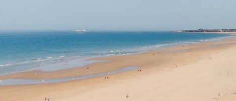 A wide sandy beach with a clear blue sea, a few people, and distant buildings.