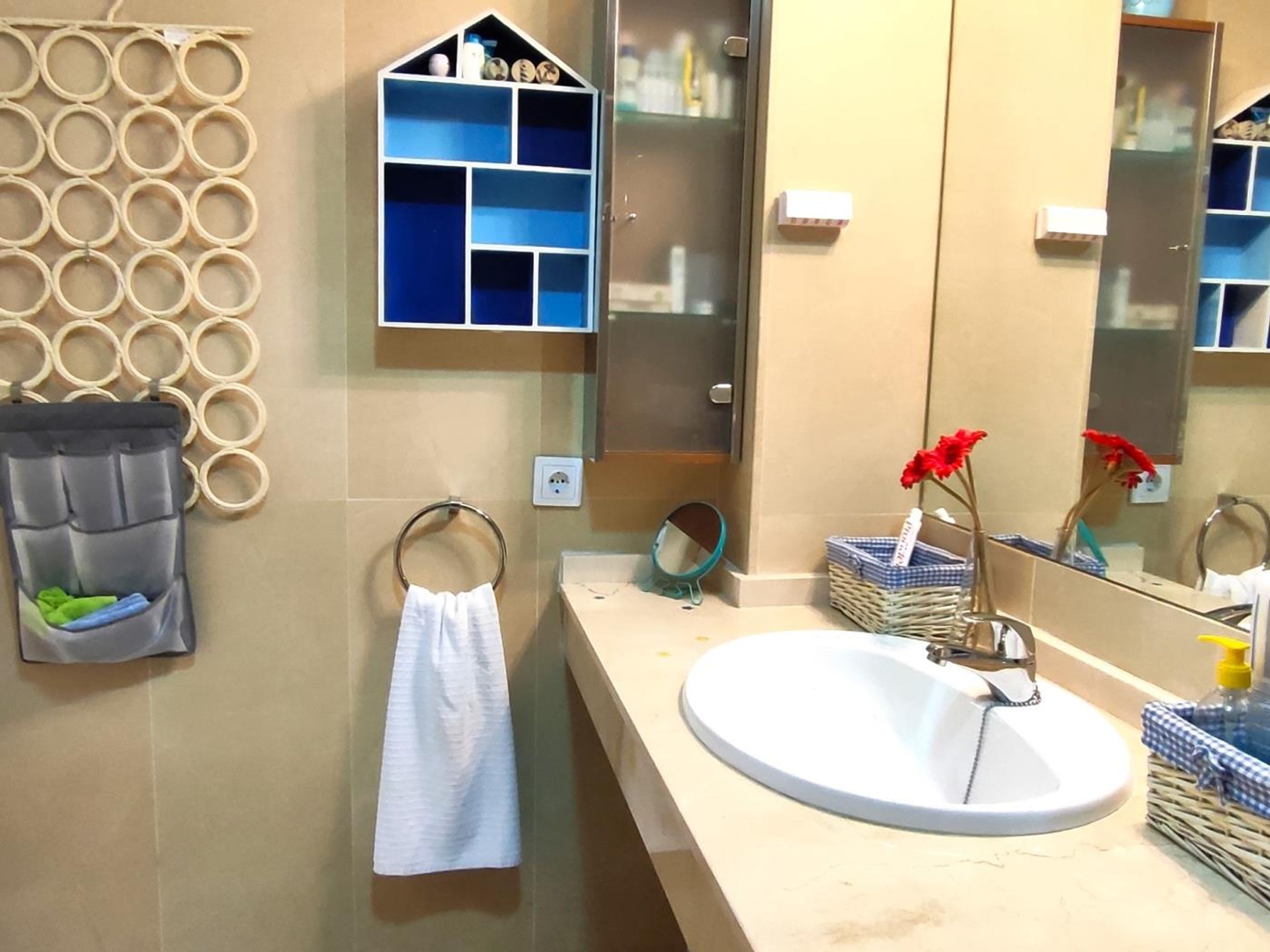 Un baño con pared de azulejos color beige, un lavamanos blanco, un espejo, un toallero, un estante pequeño con artículos de aseo y un adorno en la pared.