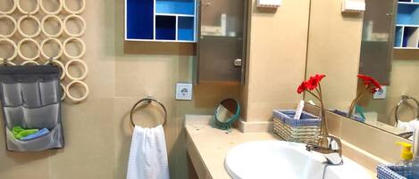 Un baño con pared de azulejos color beige, un lavamanos blanco, un espejo, un toallero, un estante pequeño con artículos de aseo y un adorno en la pared.