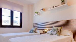 A hotel room with two beds, a large window, a bedside table with a lamp, and a shelf with books and a plant. - Coto de Sancti Petri 9 (Chiclana de la Frontera)