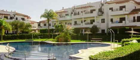 Una piscina de diseño circular, rodeada de exuberante vegetación y una hilera de edificios blancos con balcones.