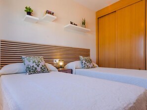 Habitación de hotel con dos camas, cabecero de madera, mesitas de noche con lámparas y estanterías de pared con plantas y libros.