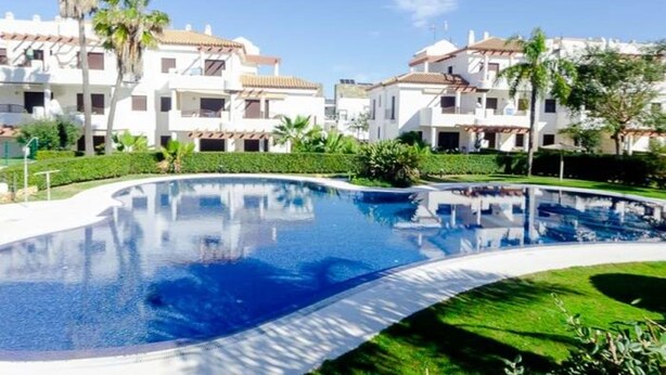 A swimming pool surrounded by white buildings with red-tiled roofs, palm trees, and well-maintained greenery.