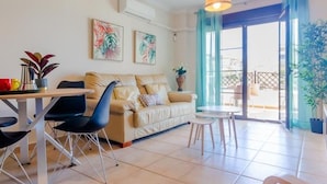 A living room with a beige sofa, a dining table with chairs, and a small table with a vase. There are two wall-mounted air conditioners and a hanging pendant light. - Coto de Sancti Petri 9 (Chiclana de la Frontera)