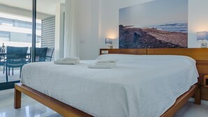 A modern hotel room with a large bed, a view of the beach through the window, and a dining area visible through the glass door. - Bah a Blanca III 18925 (El Puerto de Santa Maria)