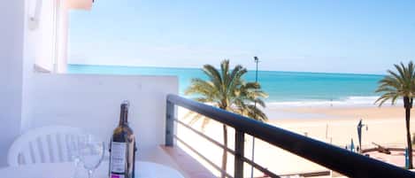 A balcony with a table, a bottle of wine, and a glass, overlooking a beach with palm trees and the ocean.