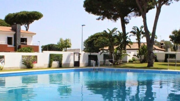 A swimming pool with clear blue water, surrounded by white walls and greenery, including trees and bushes. - Dunas 13 in Chiclana (Chiclana de la Frontera)