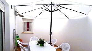 Un piccolo patio con un tavolo e sedie bianche, una pianta in vaso e un ombrellone grande.