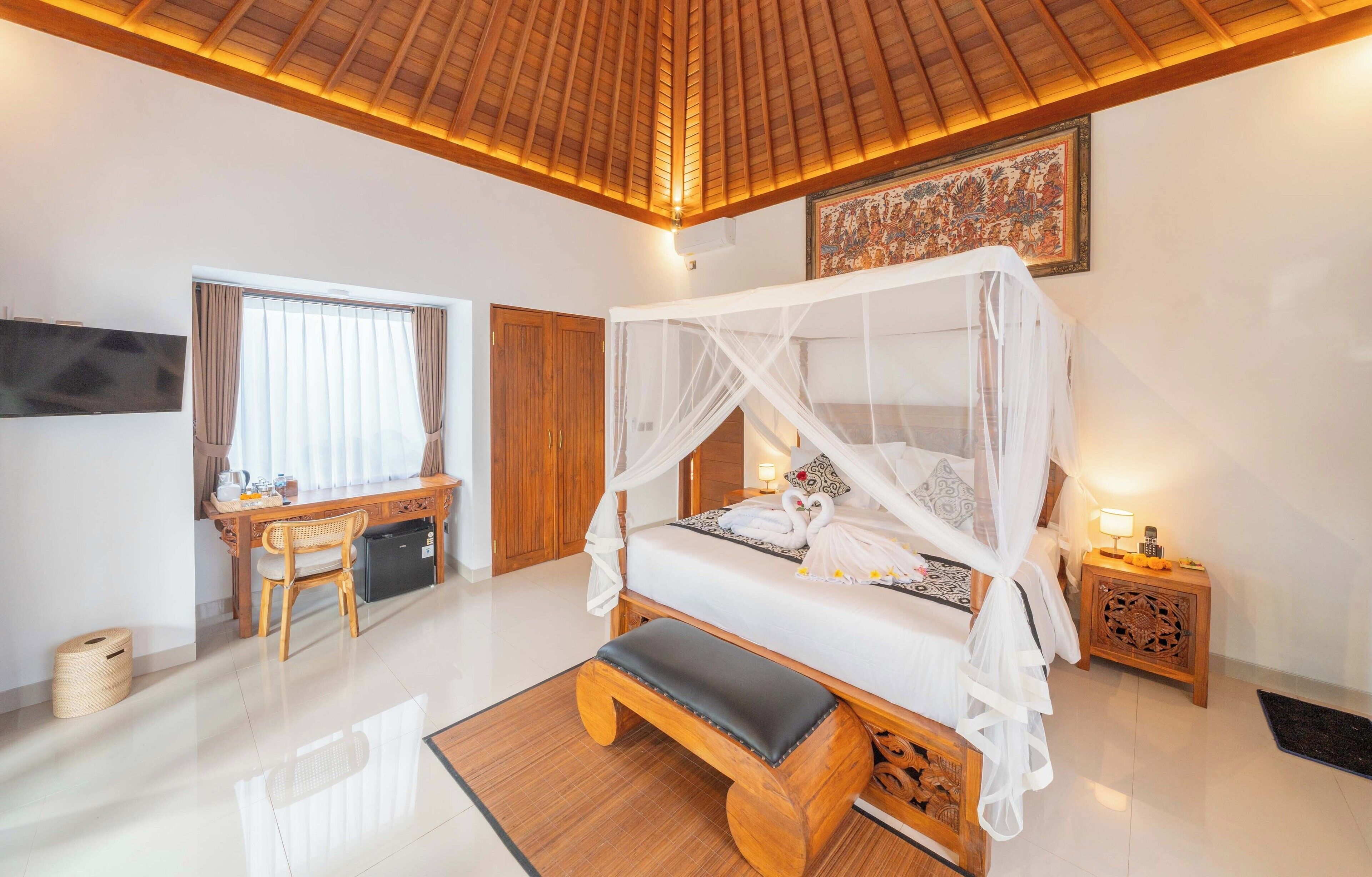 A bedroom with a four-poster bed, a wooden desk, a chair, a bedside table with a lamp, and a wall-mounted TV.